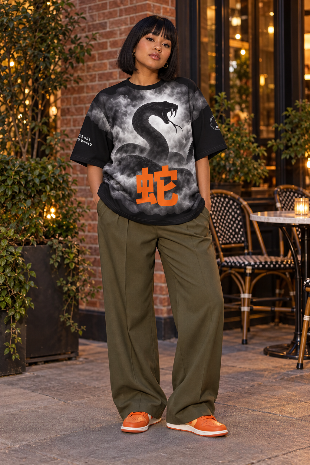 A woman stands outdoors in front of a brick building and cafe seating, wearing an oversized black graphic T-shirt with a gray snake print and orange Chinese characters, olive green wide-leg pants, and orange sneakers.