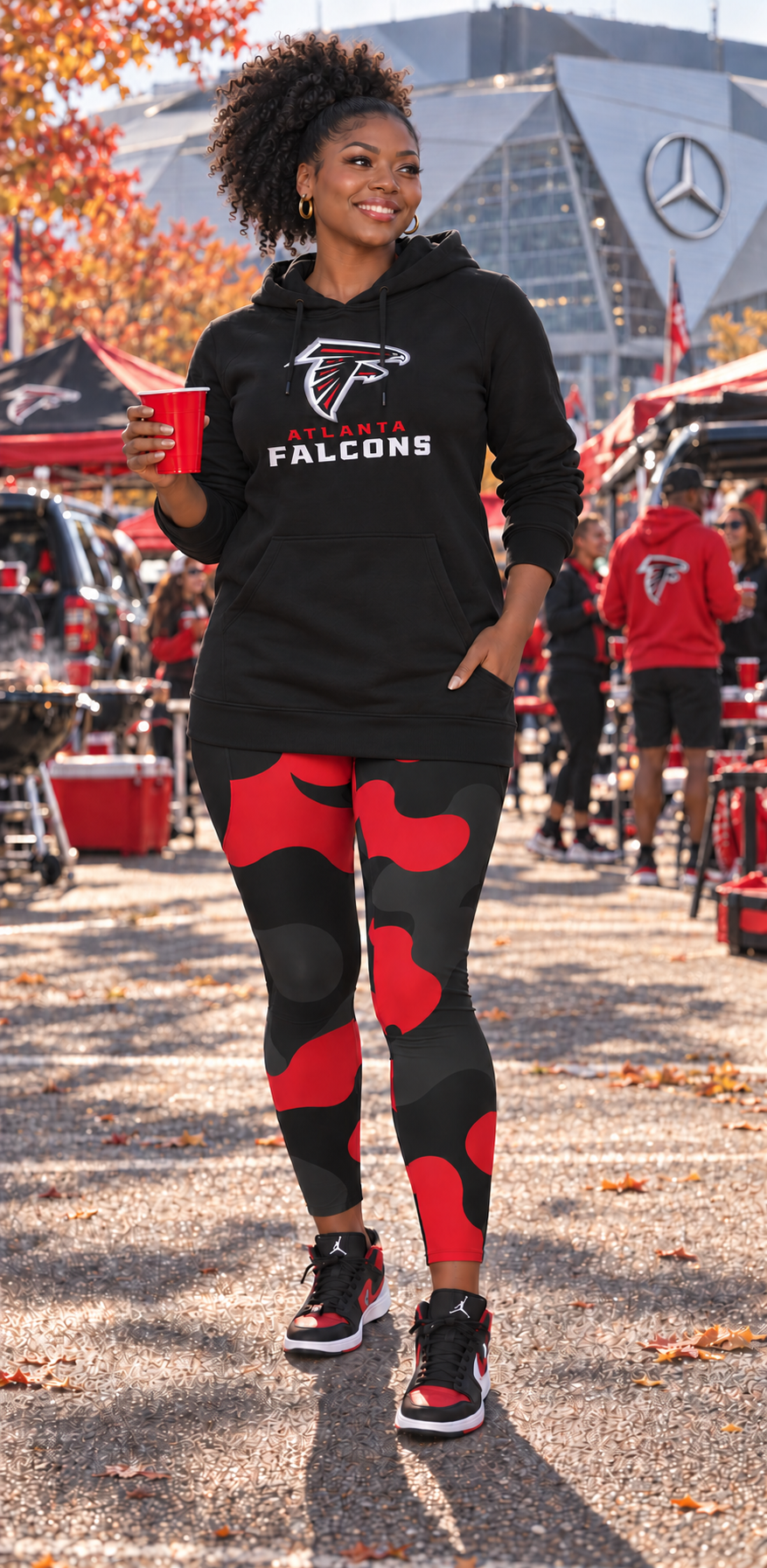 A woman stands outdoors wearing a black Atlanta Falcons hoodie and black leggings with red abstract shapes, holding a red cup. The background shows a busy outdoor event with tents, vehicles, and other people.