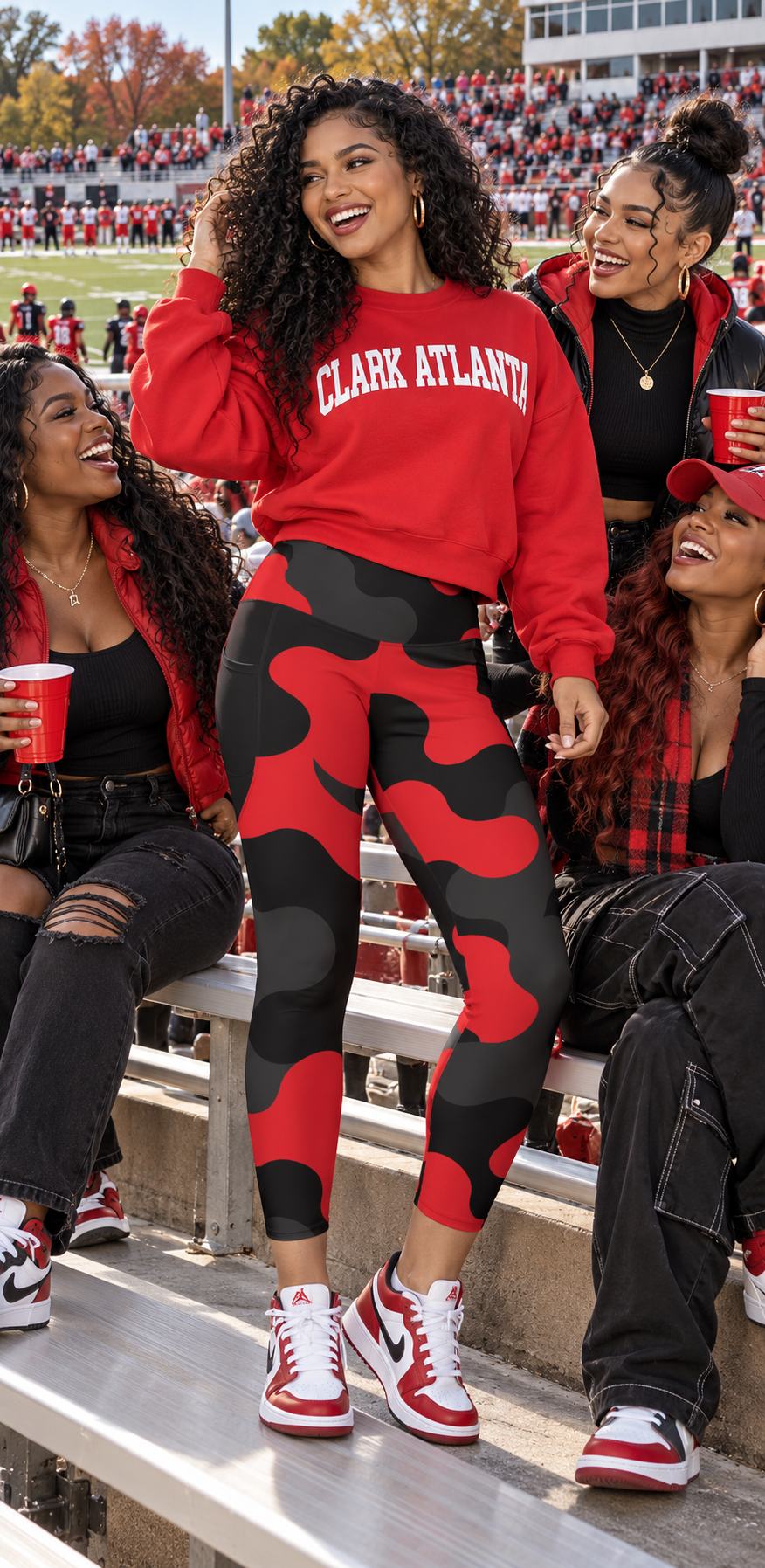 A woman sitting in stadium bleachers at a football game wearing a red 'CLARK ATLANTA' sweatshirt and red/black camouflage leggings, surrounded by friends holding red plastic cups.