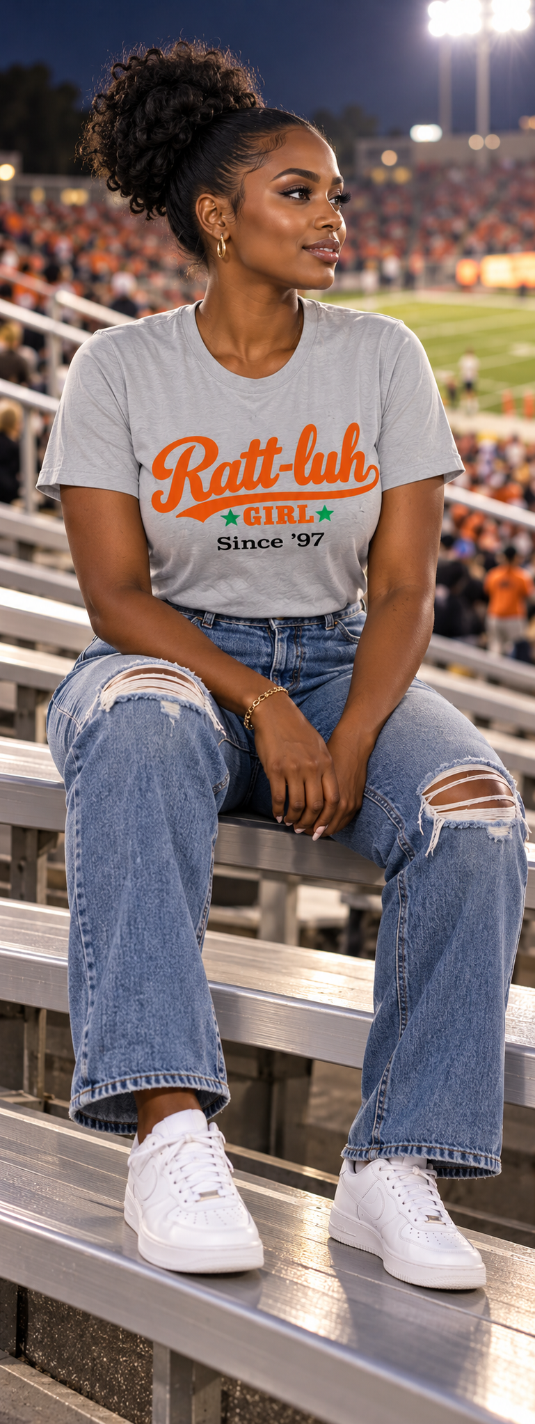 A woman sits on stadium bleachers at night wearing a light gray graphic t-shirt with orange and black text reading 'Ranit-luh GIRL Since '97', paired with distressed blue jeans and white sneakers.