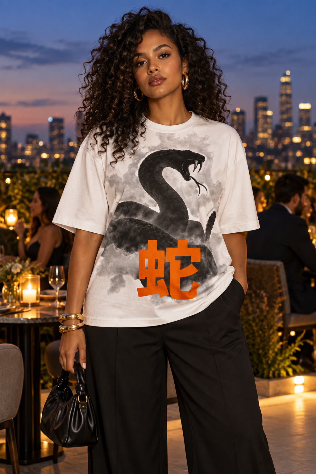 A woman stands on an outdoor rooftop at dusk wearing a white graphic T-shirt with a large black snake illustration and bold orange Chinese characters, paired with black pants. The city skyline glows in the background.