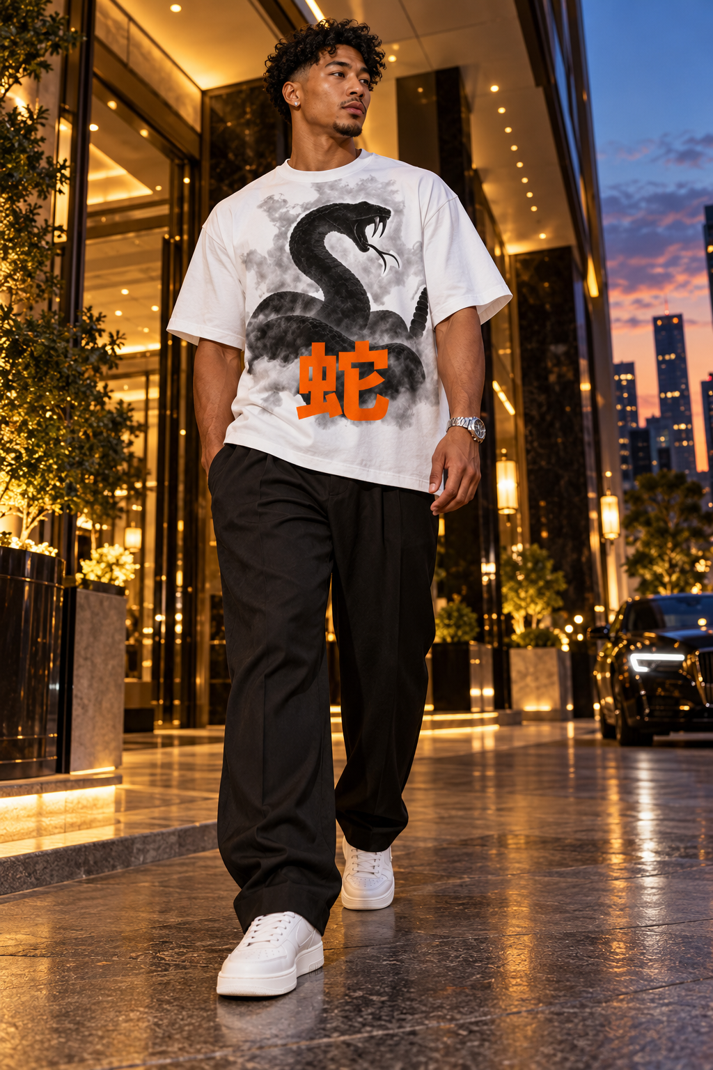 A man walking outdoors in a city setting wearing a white graphic t-shirt with a black snake print and orange Chinese characters, paired with black pants and white sneakers.