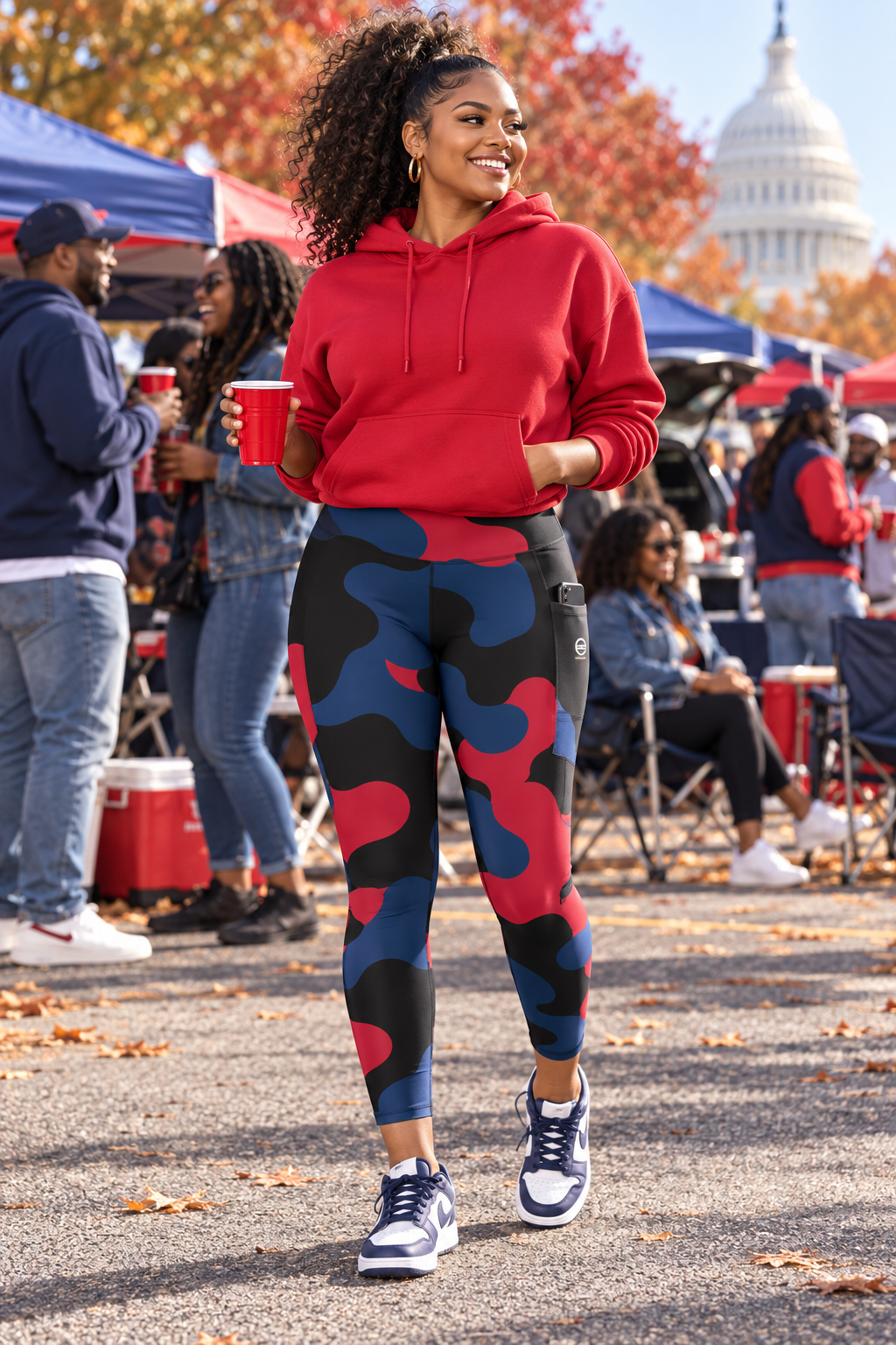 A woman walking outdoors wearing a bright red pullover hoodie and matching red, navy, and black camouflage leggings, holding red cups in a park-like setting with tents and the U.S.
